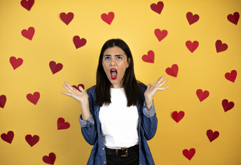 Young caucasian woman over yellow background with red hearts clueless and confused expression with arms and hands raised
