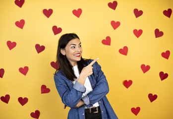 Young caucasian woman over yellow background with red hearts smiling and pointing with hand and finger to the side