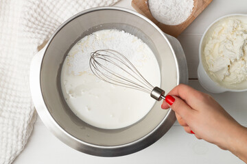 A woman's hand with a whisk over a bowl of whipping cream and powdered sugar. Flat lay.