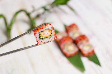 Close up of Sushi Roll piece wrapped in rice and flying fish roe in metal chopsticks with blurred background. Sushi Roll with tempura shrimp inside on bamboo leaves and plant on background. Copy space