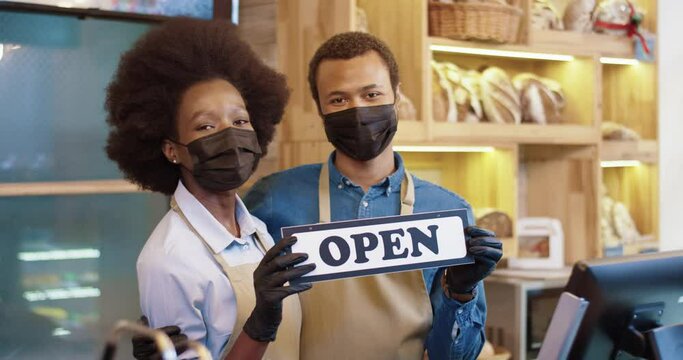Portrait of two young african american saller in apron, protective mask, and gloves holding sign "open" standing on background of shelves with bread looking at camera and smiling.