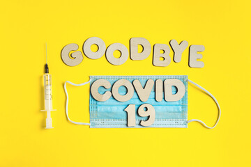 Words GOODBYE COVID 19 from wooden letters, syringe with vaccine on yellow background, and facemask. Vaccination and the end of epidemic concept.