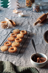 Christmas Linzer cookies filled on cooling rack, metal plate. Sandwich cookies filled with hazelnut spread, nougat cream. Hand holds cookie. . Xmas trees, candles on linen tablecloth. Natural light.