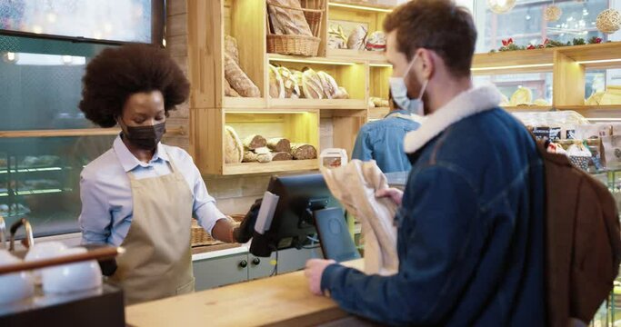 Beautiful african american saleswoman in baket shop serves client in protective mask by banking terminal following social distance. Service concept after coronavirus pandemic.