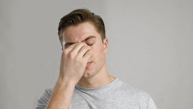 Close up portrait of young sick man rubbing his sore nose, grey studio background, slow motion