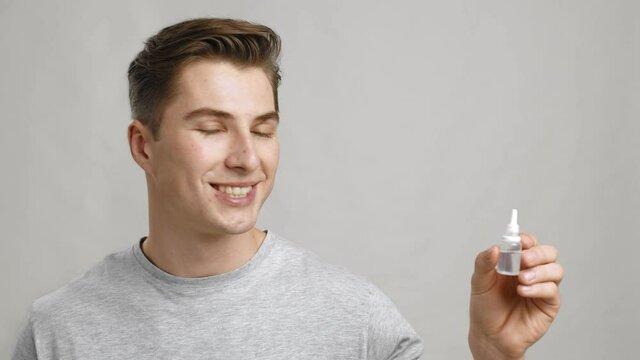 Studio portrait of young man holding bottle with medical drops and smiling to camera, grey background, slow motion