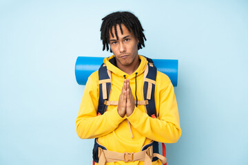 Young mountaineer african american man with a big backpack isolated on a blue background pleading