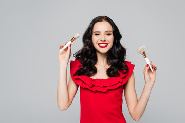 cheerful woman with red lips holding cosmetic brushes isolated on grey