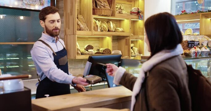 Happy baker owners work together and serves client. Woman gives goods to buyer, and man accepts payment through the terminal terminal. Contactless payment.
