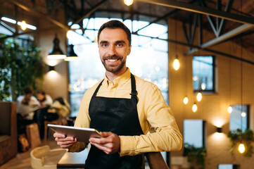 Attractive happy caucasian male waiter or barista wearing black apron stands inside a restaurant, cafe or bar, holds a menu and with friendly smile looks at the camera