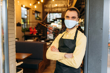 Fototapeta na wymiar A successful male waiter, barista or owner of a restaurant, cafe or bar stands at the entrance wearing a protective mask and a black work apron with crossed arms and invites clients