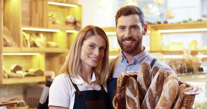Portrait of happy caucasian married couple standing in their bakery with basket of baguettes. Two family business owners are standing in their store, looking at camera smiling.