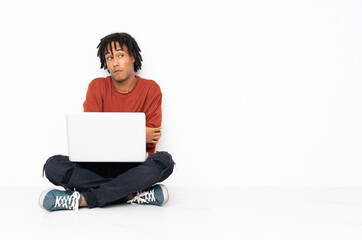 Young african american man sitting on the floor and working with his laptop making doubts gesture...