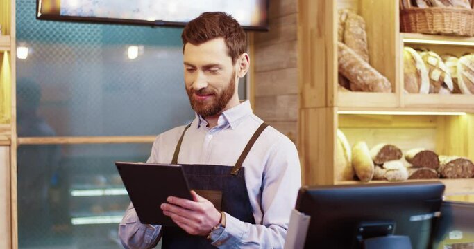 Young caucasian bakery owner standing on background of shelves with bread and communicating with client and put order in program on tablet.