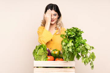 Teenager farmer girl with freshly picked vegetables in a box isolated on beige background covering a eye by hand