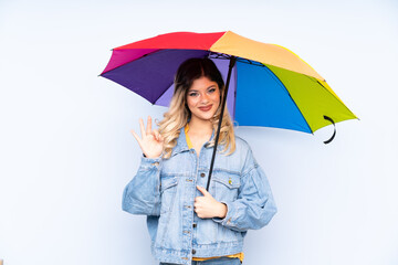 Teenager russian girl holding an umbrella isolated on blue background showing ok sign with fingers