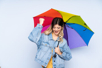 Teenager russian girl holding an umbrella isolated on blue background celebrating a victory