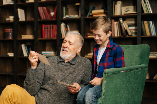 Senior bearded gray-haired grandfather with his grandson enjoy memories watching family photo album sitting on armchair and floor at home in living room with an authentic aristocratic interior.