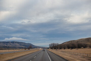 Asphalt road among steppes with yellow dry grass, blue mountains on the horizon, beautiful blue-gray autumn sky. Montana, USA, 11-23-2019