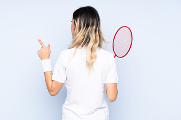 Teenager girl playing badminton isolated on blue background pointing back with the index finger