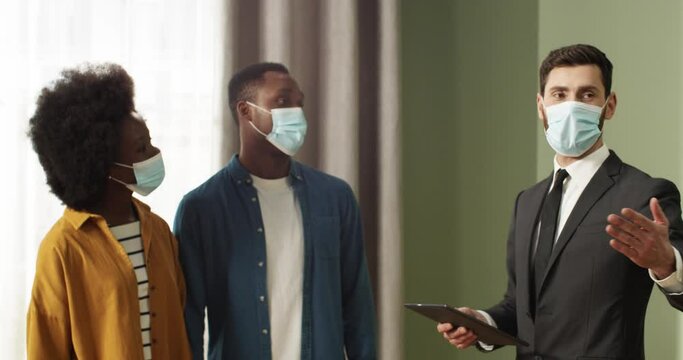 Young realtor in protective mask and suit telling about new apartment to African-American couple. Buying real estate during quarantine.