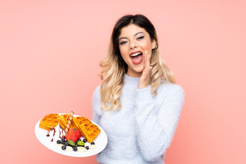 Teenager girl holding waffles on isolated pink background shouting with mouth wide open