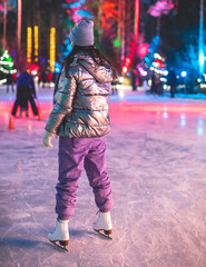 Naklejka premium Girl ice skating on the ice rink arena with happy people in the background, concept of ice skating in winter, winter activities, holiday christmas time, with new year decoration and illumination