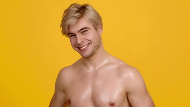 Playful shirtless blonde guy shooting to camera with hand and smiling, winking to camera, orange studio background