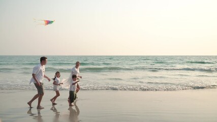 Happy Asian family on summer holiday vacation. Parents with little child boy and girl run and play kite together on tropical beach at summer sunset. Family enjoy and having fun with outdoor activities