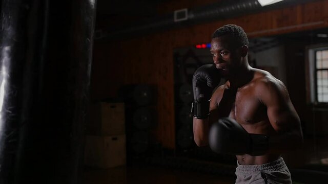 Black athletic shirtless young man boxer training punching on the punching bag with an effort in the gym