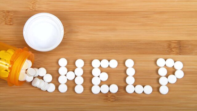 HD video zooming on bottle of pain pills spilled on wood table spelling the word HELP.  Addiction and opioid epidemic theme zooming in on the word help
