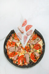 Directly above of Italian meat black dough Pizza on white marble background with ingredients: tomato slices, ham, bacon, salami, pork and beef meat. Flying food concept. pizza served with pesto sauce.