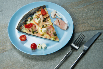 Black dough pizza cut with ingredients on blue plate. Knife and fork near the dish on stone anthracite background. Piece of pizza with cherry tomatoes, mushrooms, bacon, feta cheese, pesto sauce
