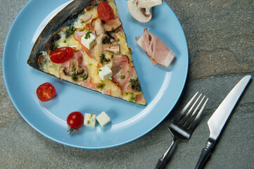 Slice of black dough pizza with ingredients on blue plate. Knife and fork near the dish on stone anthracite background. Piece of pizza with cherry tomatoes, mushrooms, bacon, feta cheese, pesto sauce