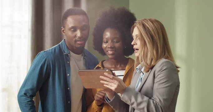 Professional female real estate agent showing young african american couple apartment plan using electronic tablet. Home buying concept.