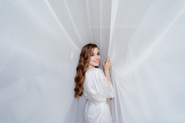 beautiful young woman with long hair in a white robe stands under a curtain