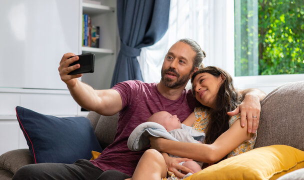 Caucasian man using smartphone taking selfie or home video of his family wife and adorable newborn baby son in living room. Father and mother relax and enjoy with infant child boy together at home.