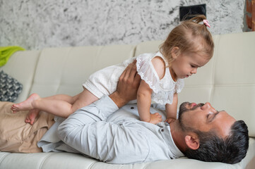 A Caucasian man lies on the couch and holds a little daughter above him. Caring loving dad raised the child up. Happy fatherhood.
