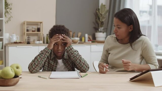 Medium close-up of guilty African school boy holding head with hands, sitting by desk at home, annoyed Mixed-Race woman scolding son for not doing homework correctly