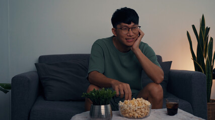 Asian man watching a living room series while eating a popcorn at night.