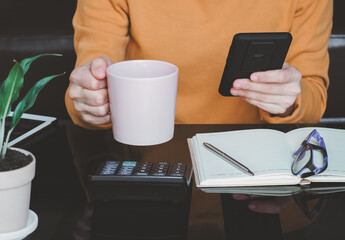 asian young woman holding coffee cup and using smartphone shopping online-calculate home salary and expenses. concept saving money investment.