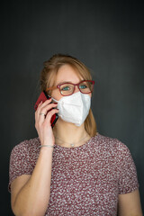 Junge blonde Frau mit Brille telefoniert mit FFP2 Schutzmaske - isoliert vor dunkelgrauem Hintergrund mit Textfreiraum- rote Accessoires