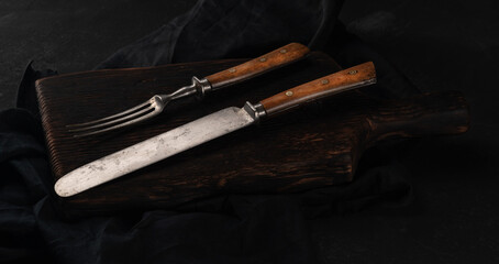 Vintage knife and fork on kitchen board with napkin, black background