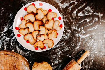 Cookies in shape of heart for the Saint Valentine's Day. Dough, flour, baking pan, round wooden cutting board and rolling pin on the table. Cookies on the plate.