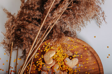 Heart-shaped cookies on round wooden cutting board and dries pampas grass. Saint Valentine's cookies in shape of heart and golden sparkles. Golden glitters in shape of heart on white background.