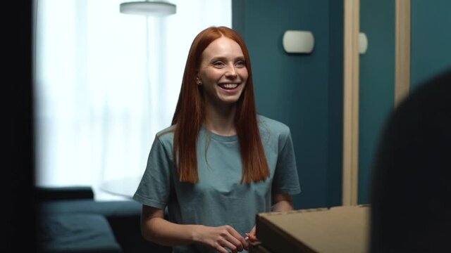 Smiling redhead woman listening delivery man, laughing and receiving boxes with hot pizza and going at home. Back view of courier delivering food on doorstep at apartment. Tracking shot in slow motion