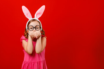 Happy easter. Portrait of a cheerful little girl in bunny ears and glasses on a red background. The child has fun and pretends to be a rabbit.