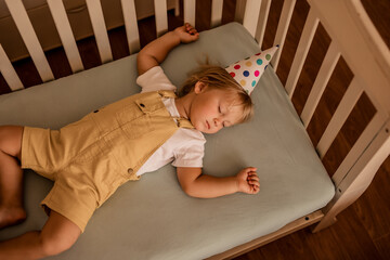 A two-year-old boy in a festive paper hat sleeps in his bed after the holiday