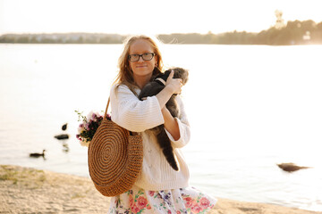 Young blonde girl with glasses holds a cat breed Scottish Straight. Walk with pet near the lake, river at sunset on the beach