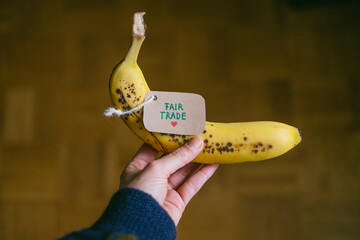 One hand holds a banana with a label that says Fair Trade. Consume fair trade goods and conscious...